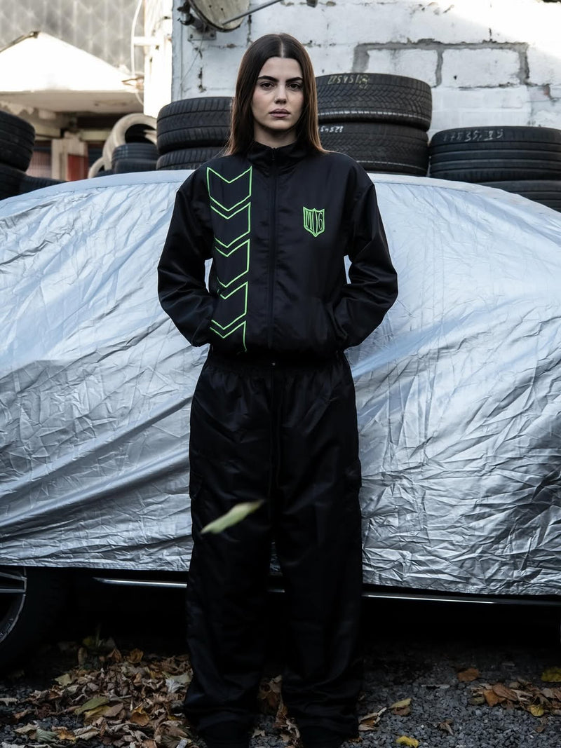 Person wearing a black jumpsuit with green accents standing in front of a covered vehicle.