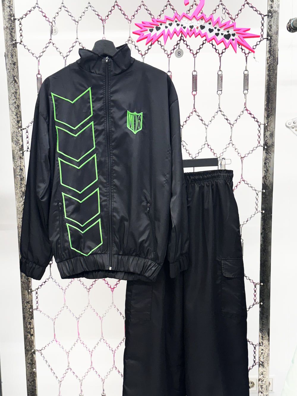 Black jacket with green accents and black pants on a hanger against a textured white wall.