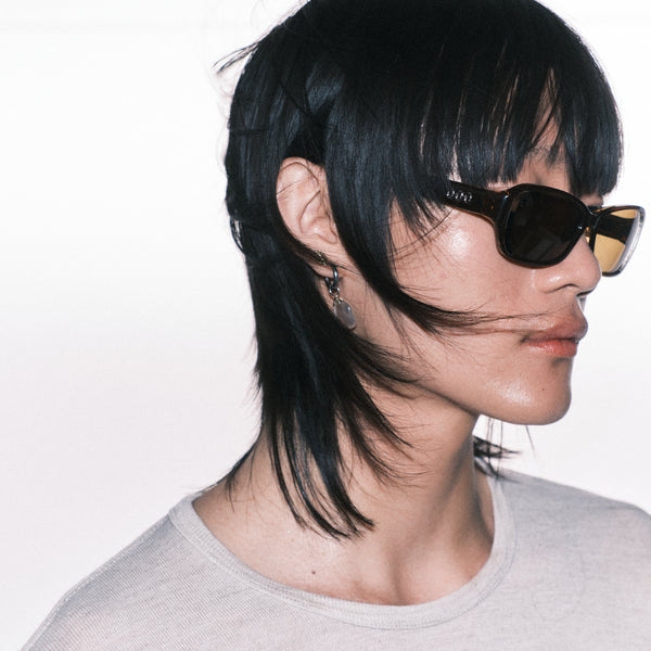 Person with black hair styled in a braid, wearing sunglasses and a gray shirt on a white background
