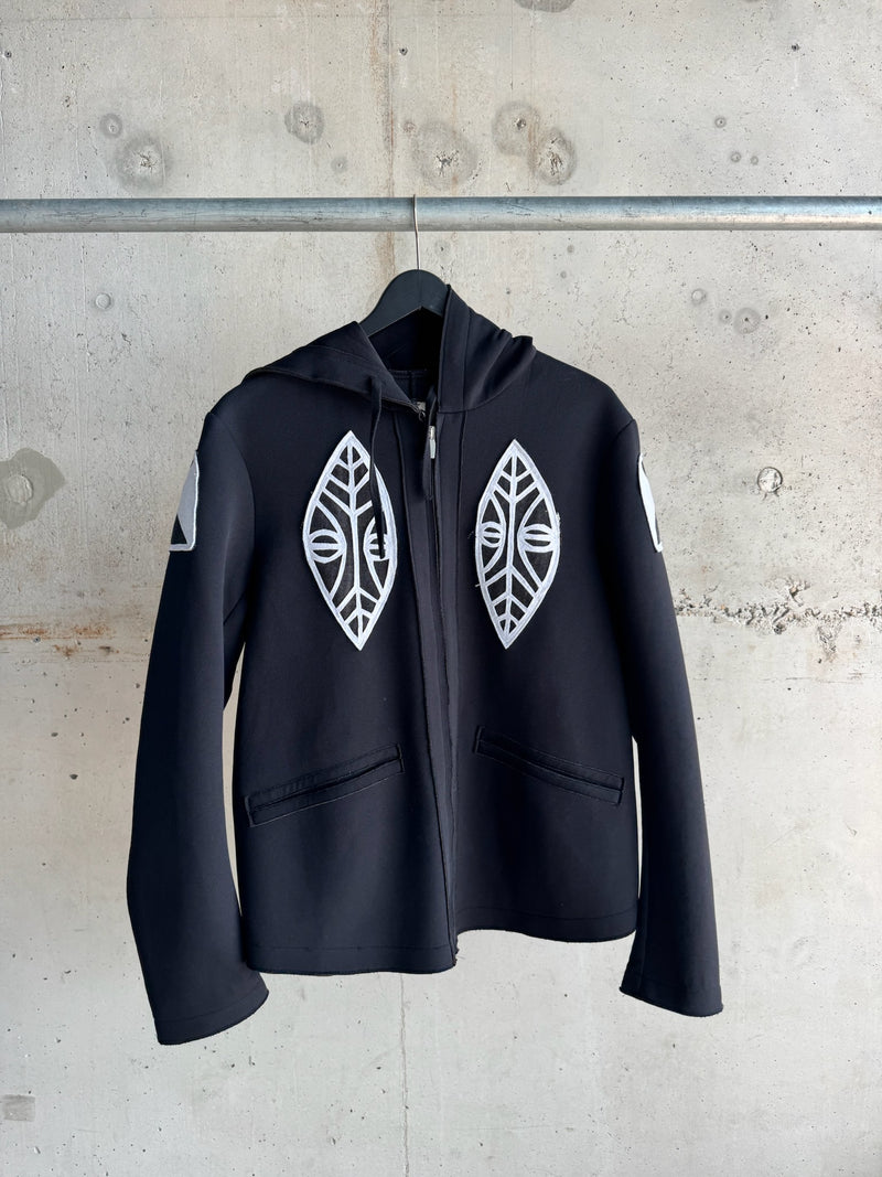 Black zip-up hoodie with bold white tribal-style face and leaf appliqués on the chest and sleeves, displayed on a hanger against a raw concrete wall
