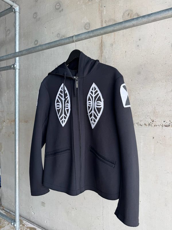 Angled view of a black hoodie with front zip closure, featuring white tribal-inspired face and leaf motifs on the chest and a geometric patch on the sleeve, hung against a concrete wall