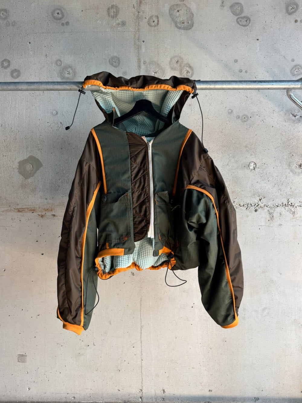 Front view of an oversized green and brown hooded bomber jacket with orange trim, front zipper, drawstring hood and hem, and quilted inner lining, displayed on a hanger against a concrete wall background.