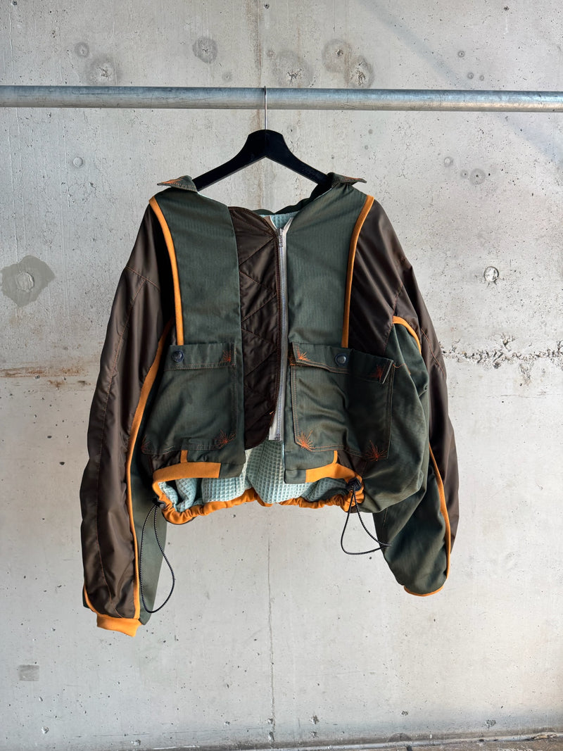 Oversized green and brown patchwork bomber jacket with orange piping, front zipper, and drawstring hem, featuring utility-style pockets and a textured inner lining, displayed on a black hanger against a concrete wall