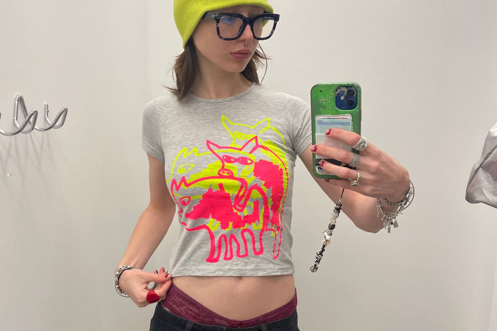 Person taking a mirror selfie wearing a gray t-shirt with a colorful graphic, black cargo pants, and a yellow beanie.