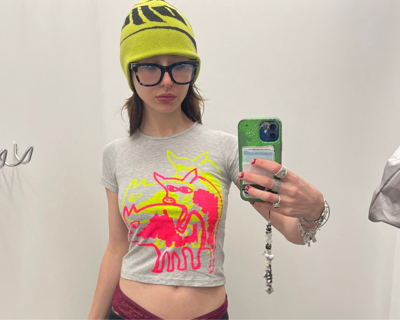 Person taking a mirror selfie wearing a gray t-shirt with a colorful graphic, black cargo pants, and a yellow beanie.