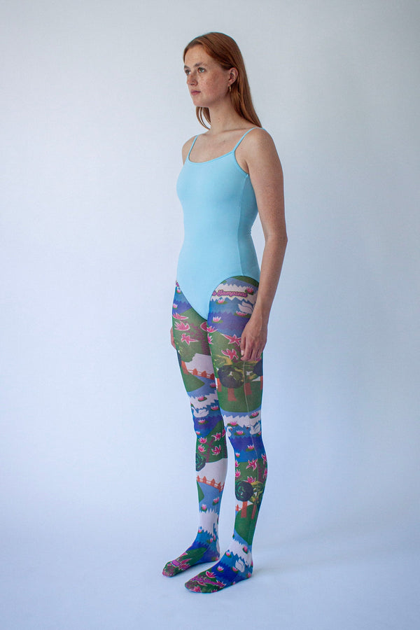 Side profile of a female model wearing a light blue camisole-style bodysuit and colorful scenic print leggings, standing against a plain white studio background.