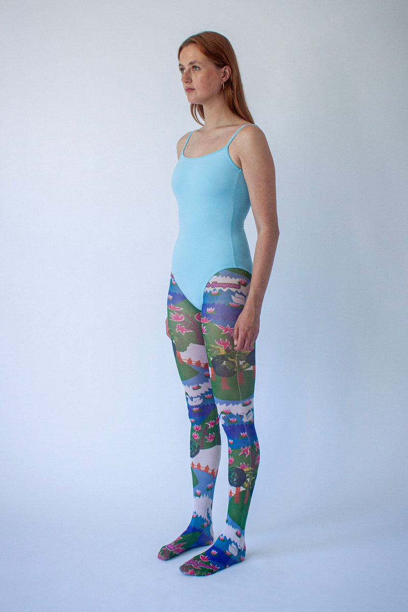 Side profile of a female model wearing a light blue camisole-style bodysuit and colorful scenic print leggings, standing against a plain white studio background.