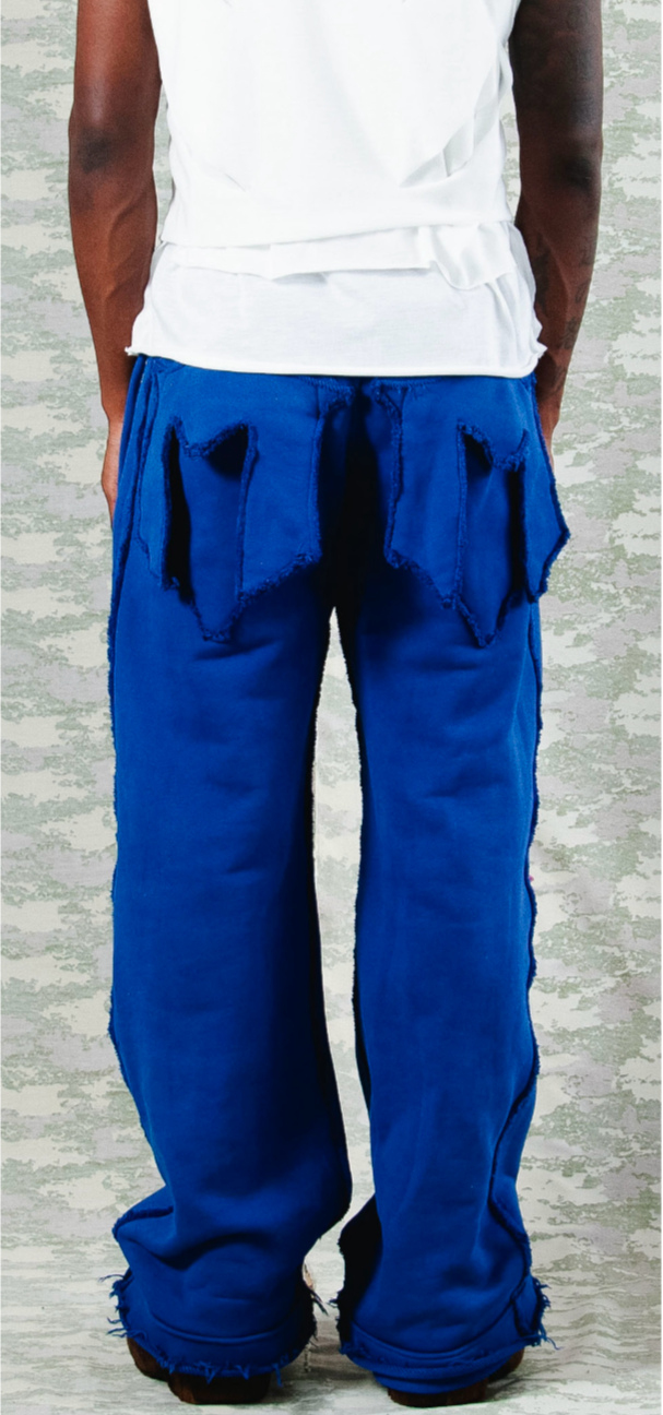 Back view of cobalt blue wide-leg pants with oversized raw-edge back pockets and frayed hems, styled with a layered white sleeveless top – urban fashion look against camo-print backdrop