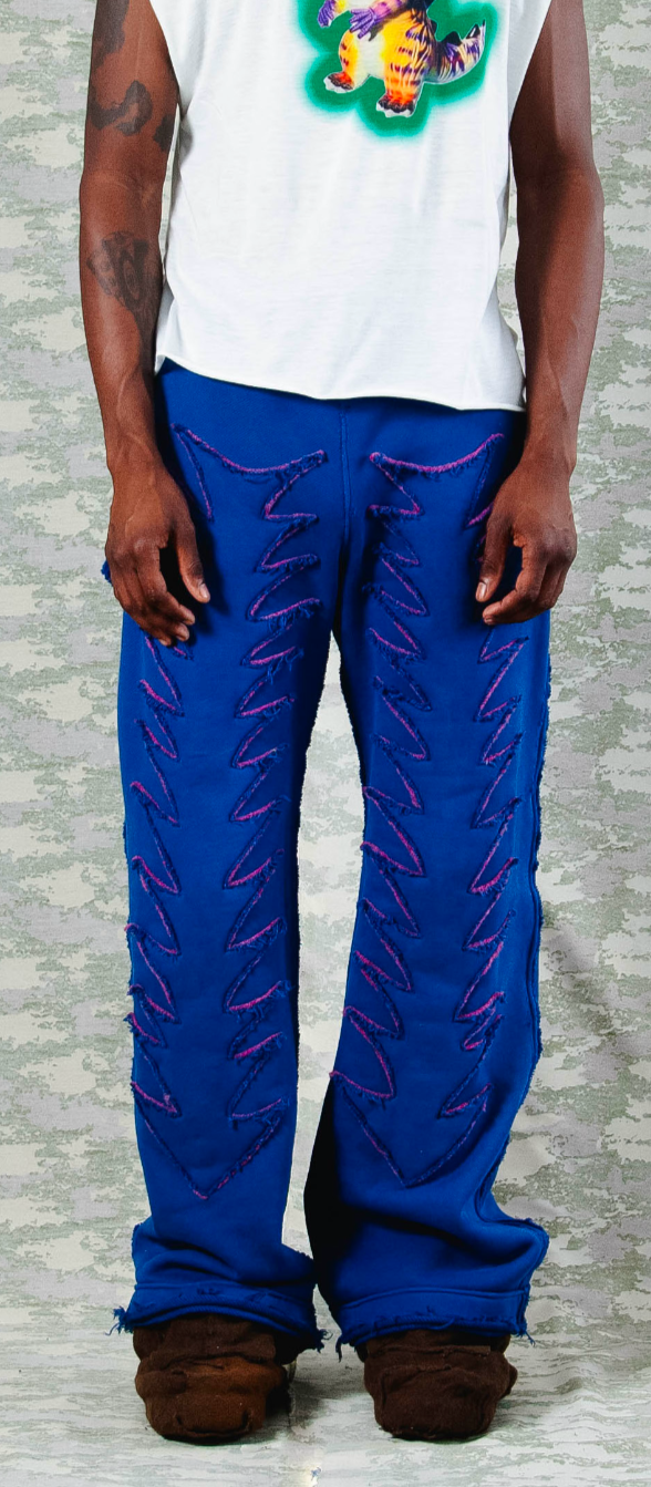 Front view of electric blue wide-leg pants with bold purple zigzag stitching and raw hems, styled with white sleeveless tee featuring a vibrant dinosaur graphic – edgy streetwear look on camo backdrop