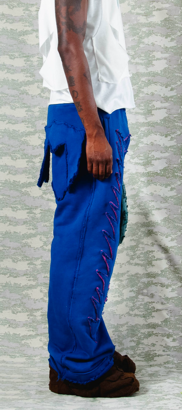 Side view of electric blue wide-leg pants with exaggerated purple zigzag stitching and oversized raw-edged pocket, styled with layered white sleeveless top – camo backdrop enhances streetwear silhouette