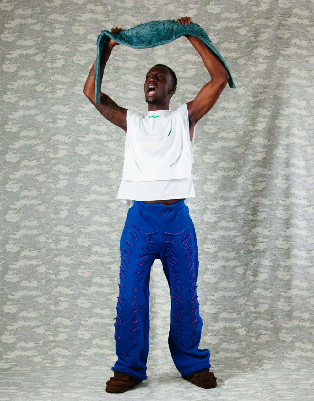 Full-body shot of model lifting teal textured fabric above head, wearing a layered white sleeveless top and electric blue pants with exaggerated purple zigzag stitching – posed energetically against a camo-patterned backdrop