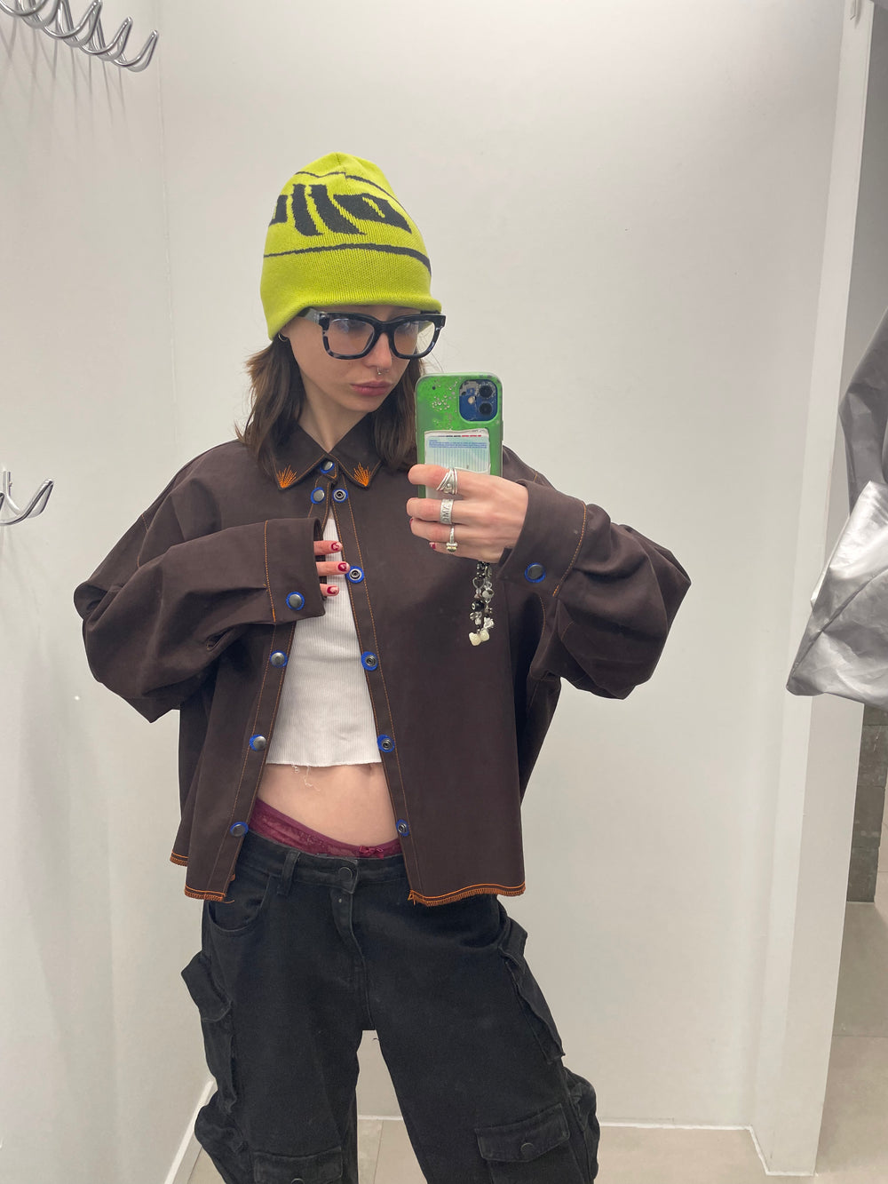 Person wearing a brown jacket, green beanie, and black pants taking a mirror selfie.