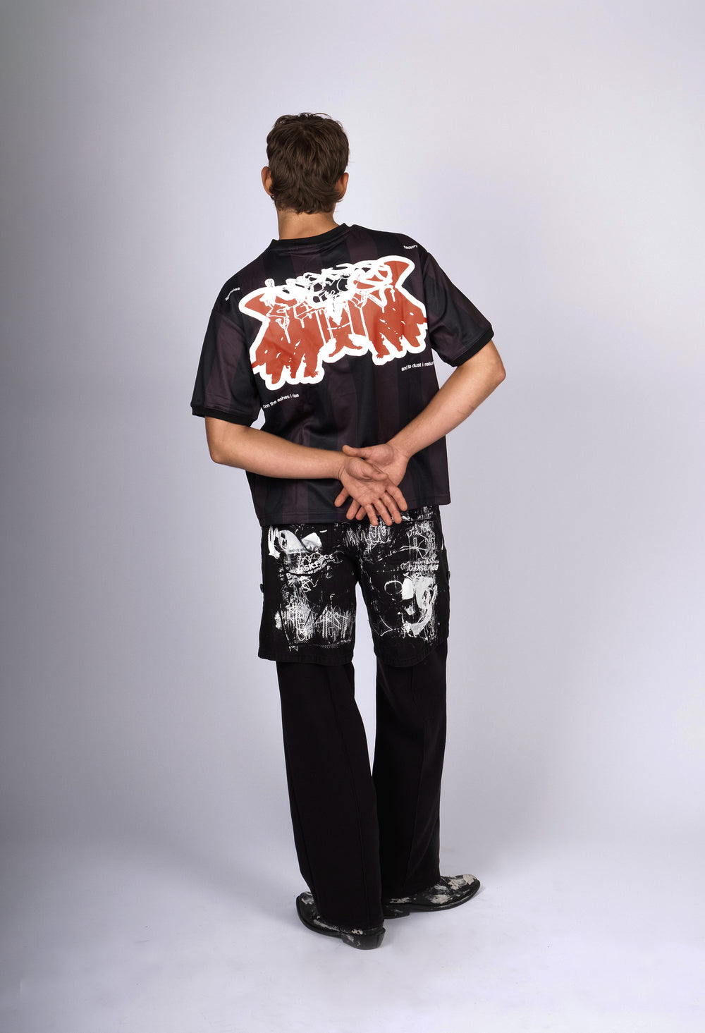 Model wearing a black and dark red striped T-shirt with a bold red and white graphic on the back, featuring abstract wing-like shapes and the text 'from the ashes I rise and to dust I return.' Paired with black pants splattered with white paint details, the outfit combines edgy streetwear with artistic elements for a contemporary, expressive look