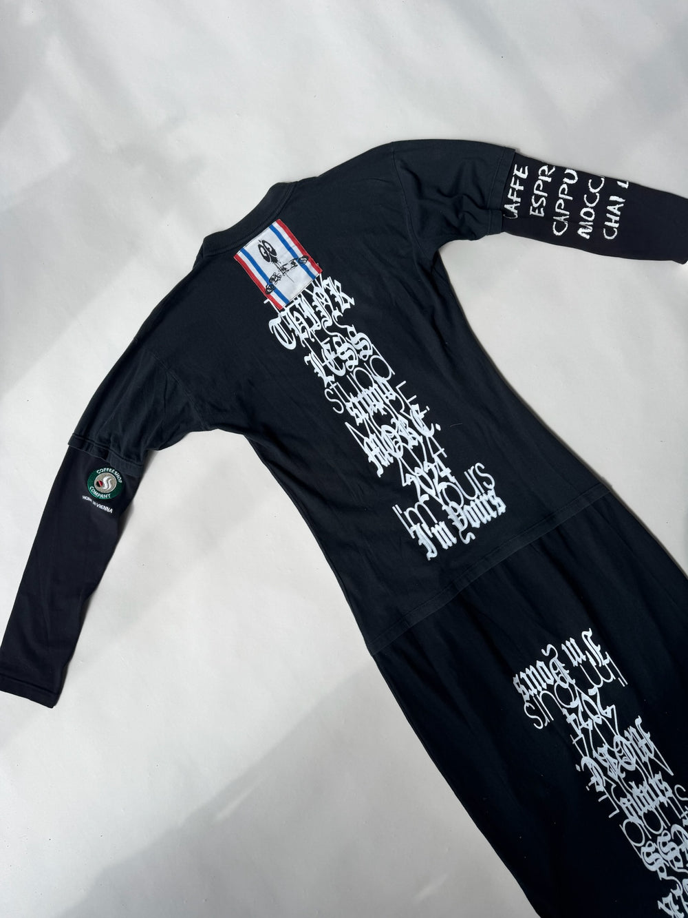 Flat lay of black upcycled maxi dress with white gothic script reading ‘Think Less Stupid More Sex 2024 I’m Yours’, repurposed sleeve patches, and a red-white-blue stitched ribbon detail at the neckline