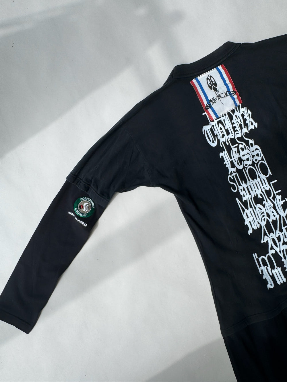 Back view of black graphic dress by GATTO des F*CKDOWN featuring white gothic typography, red-white-blue patch near collar, and embroidered Coffee Shop Company logo on left sleeve