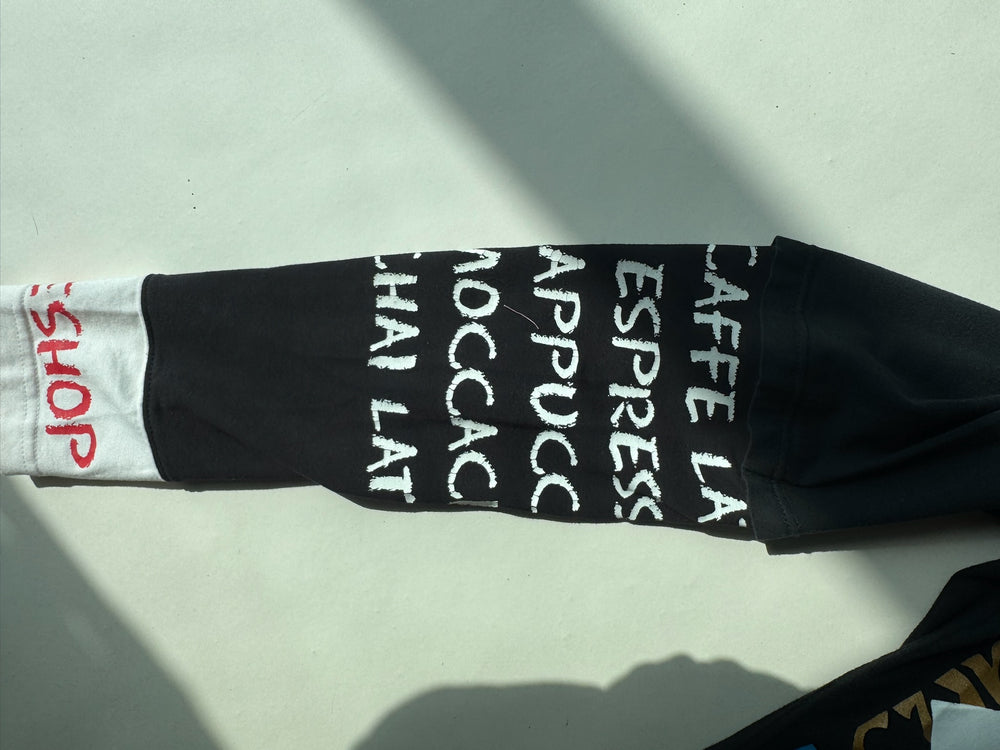Close-up of black sleeve with white handwritten-style coffee-related text including ‘CAFFE LATTE’, ‘ESPRESSO’, ‘CAPPUCCINO’, ‘MOCCACCINO’, and ‘CHAI LATTE’, ending in a white cuff with red ‘COFFEE SHOP’ detai