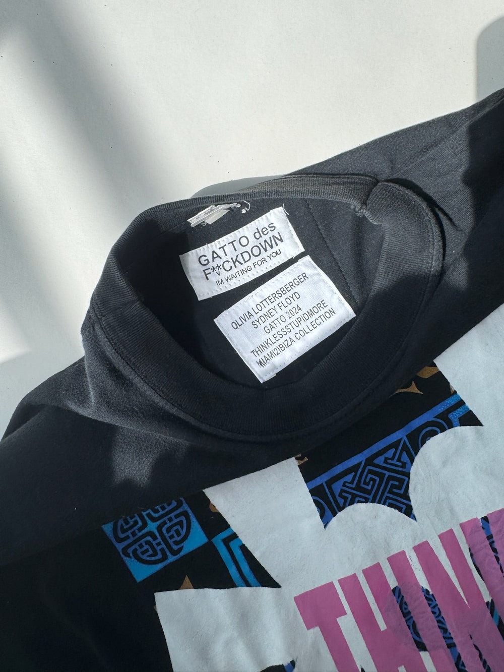 Close-up of black dress neckline showing GATTO des F*CKDOWN label by Olivia Lottersberger, part of the STUPID MORE SEX Studio Collection, featuring white printed tags inside the collar
