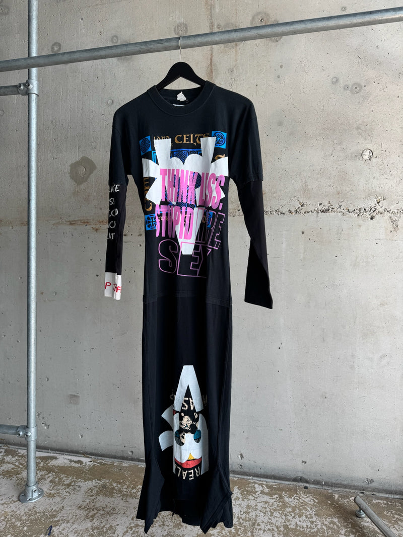 Black upcycled maxi dress with bold graphic prints, text ‘Think Less Stupid More Sex’, long sleeves, and mixed pop culture visuals, hanging against industrial concrete wall