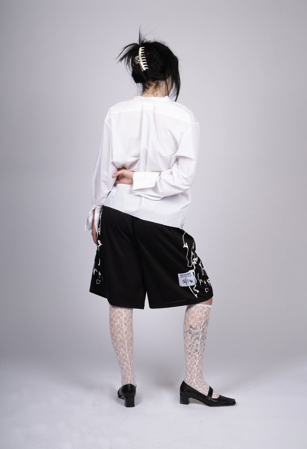 Model wearing black athletic shorts with white abstract line designs along the sides and a branded patch, styled with a white oversized shirt, patterned lace stockings, and black loafers. The outfit combines a mix of sporty and chic elements for a unique, contemporary look