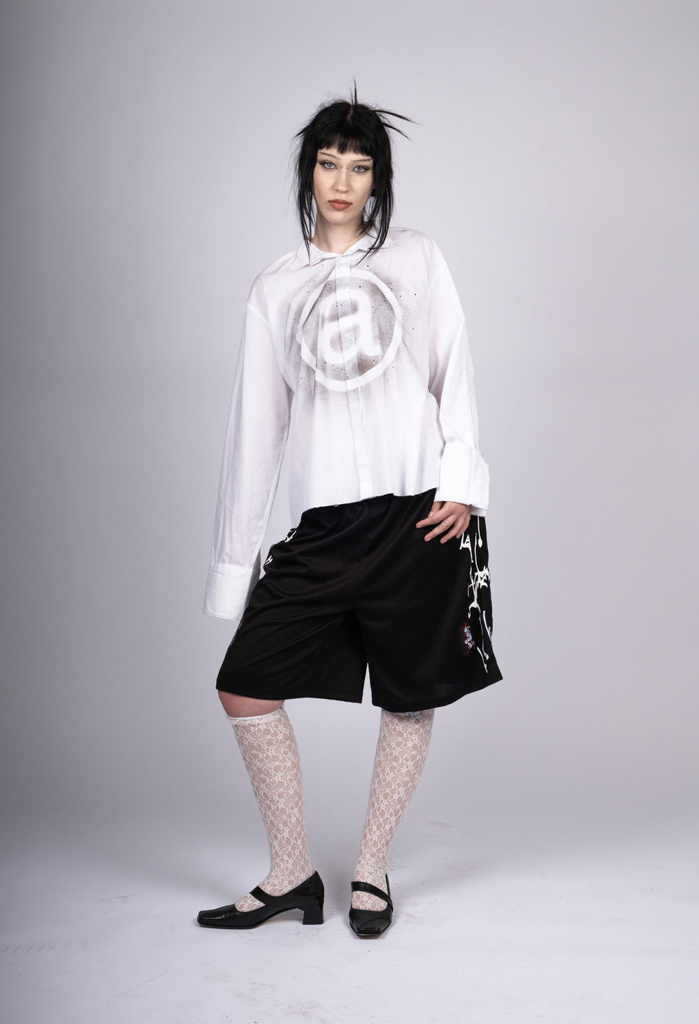 Model wearing black athletic shorts with white abstract designs on the sides, paired with a loose white shirt featuring a large '@' symbol graphic, patterned lace stockings, and black Mary Jane shoes. The outfit combines edgy streetwear with playful, vintage-inspired elements, creating a bold, contemporary look.