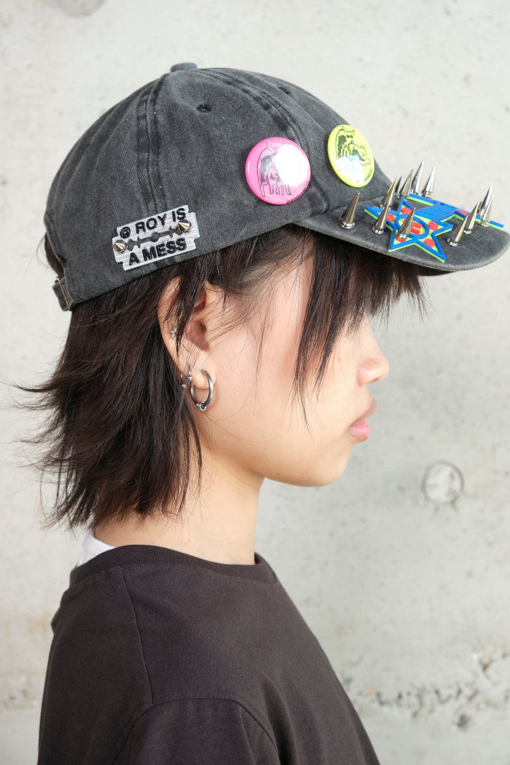 Close-up portrait of a young person wearing a black spiked cap with colorful 3D graphic details and @royisamess pins, styled with a black t-shirt against a concrete wall backdrop.