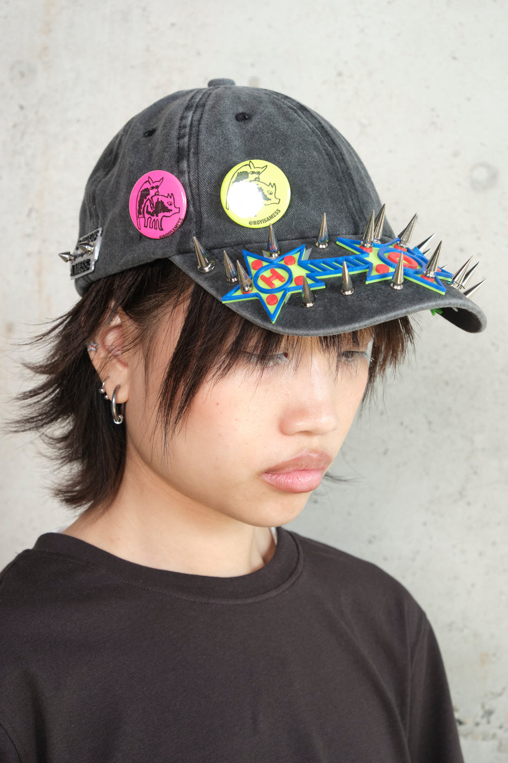 Side profile of a person wearing a distressed black cap customized with neon-colored pins, metallic spikes, and bold graphic embellishments in blue, red, and green. The cap features two badges—one pink and one yellow—with a mirrored animal illustration and the handle @ROYISAMESS. The person has layered earrings and wears a black top, captured against a concrete background