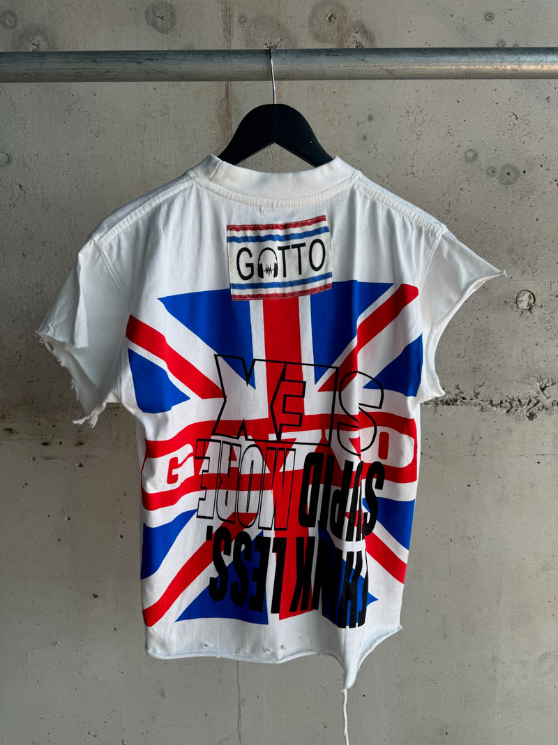 Back view of a white distressed T-shirt with inverted bold black text “THINK LESS STUPID MORE SEX,” Union Jack graphic, and a sewn-in “GATTO” label, hanging on a black hanger against a concrete wall