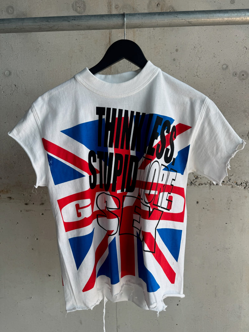 White distressed T-shirt featuring bold black text “THINK LESS STUPID MORE SEX” over a Union Jack flag graphic, hanging on a black hanger against a concrete wall.