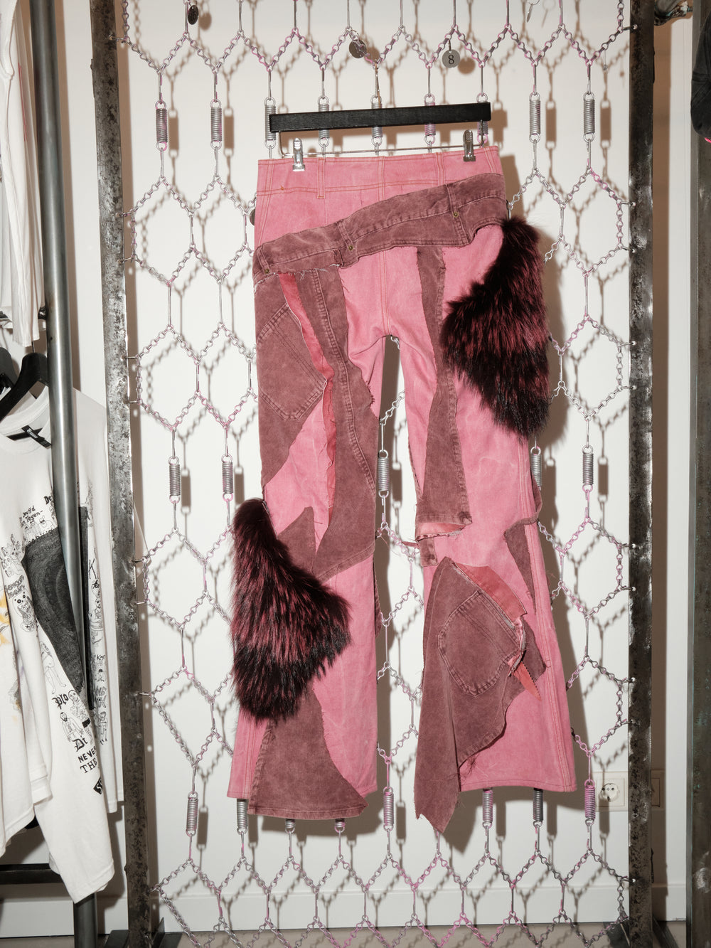 A pair of pink patchwork pants hanging on a black hanger against a chain-link patterned backdrop. The pants feature asymmetrical brown suede panels, frayed seams, butterfly graphics, and large dark faux-fur accents on the sides and lower legs, giving them a deconstructed, avant-garde look.