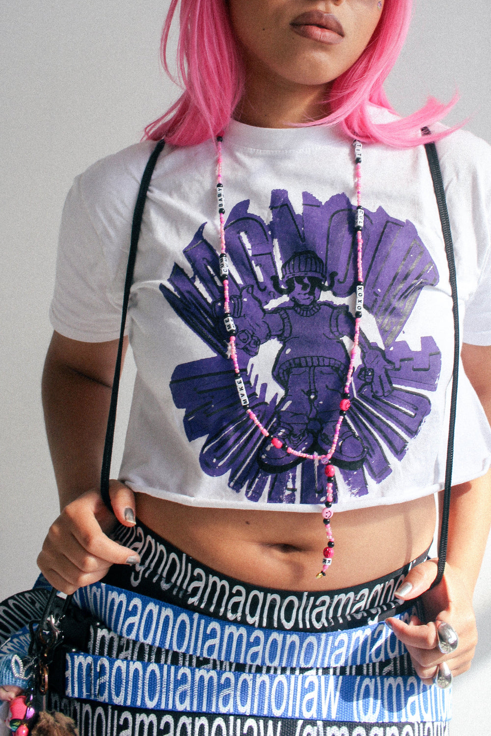Model wearing a Magnolia cropped graphic T-shirt with purple print, styled with layered Magnolia logo straps worn as belts and accessories.