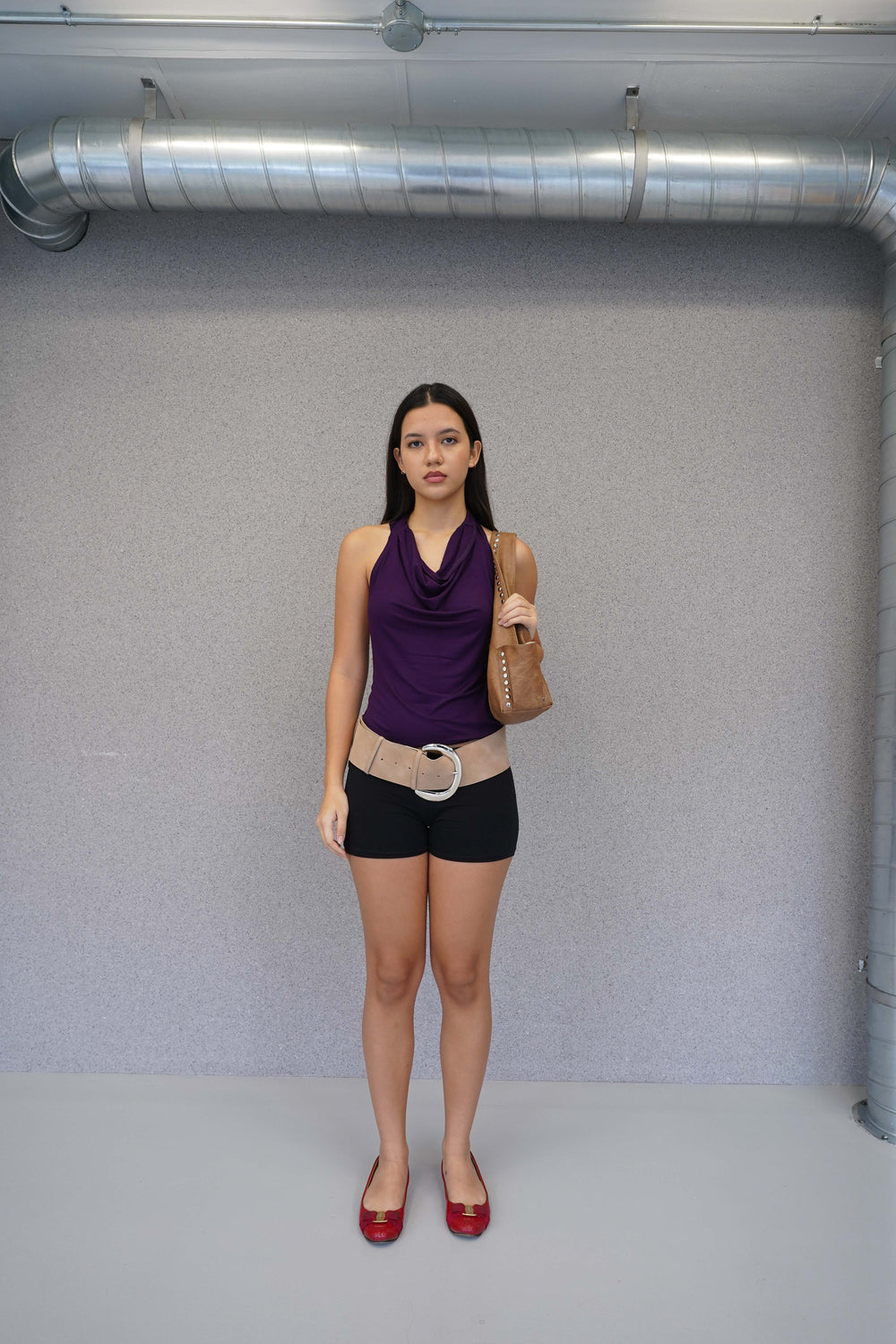 Woman in a purple top and black shorts standing against a plain wall with visible pipes.