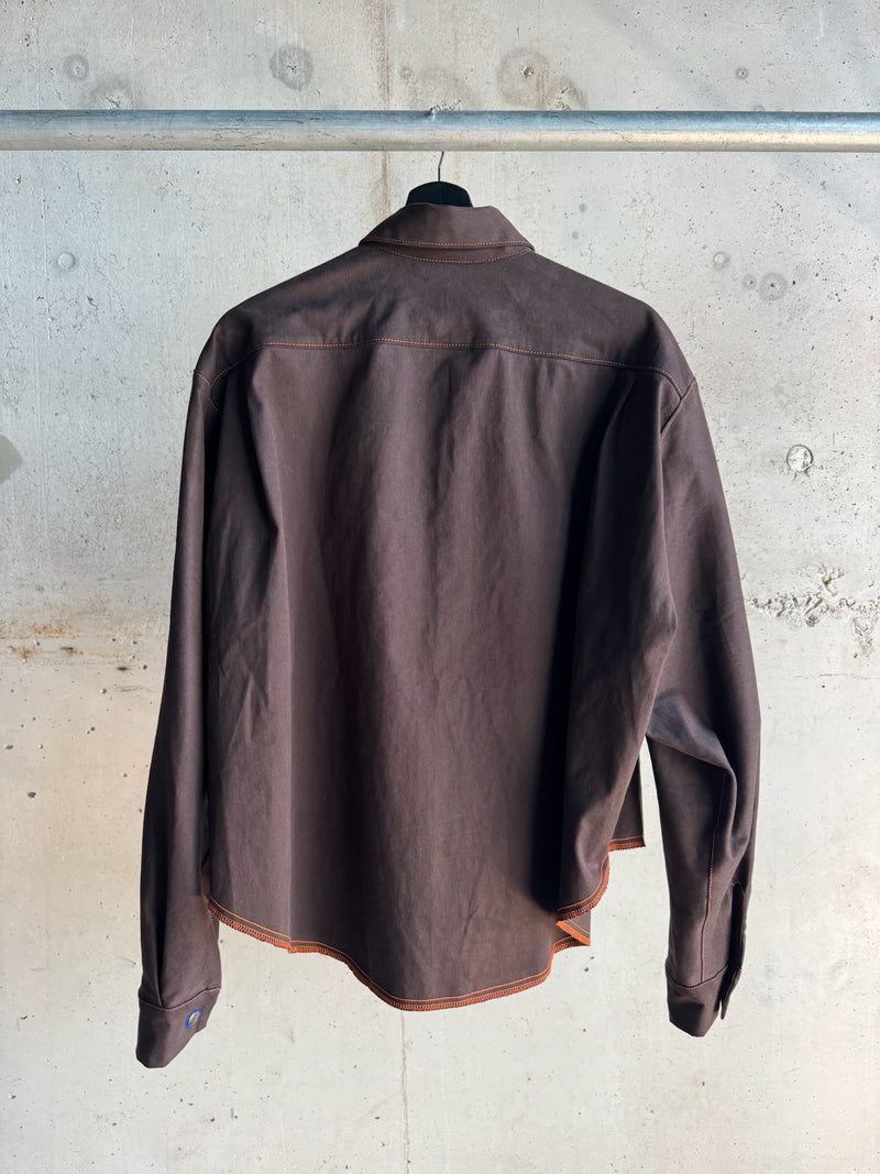 Back view of a dark brown long-sleeve shirt with orange contrast stitching and a curved hem, displayed against a raw concrete wall backdrop.