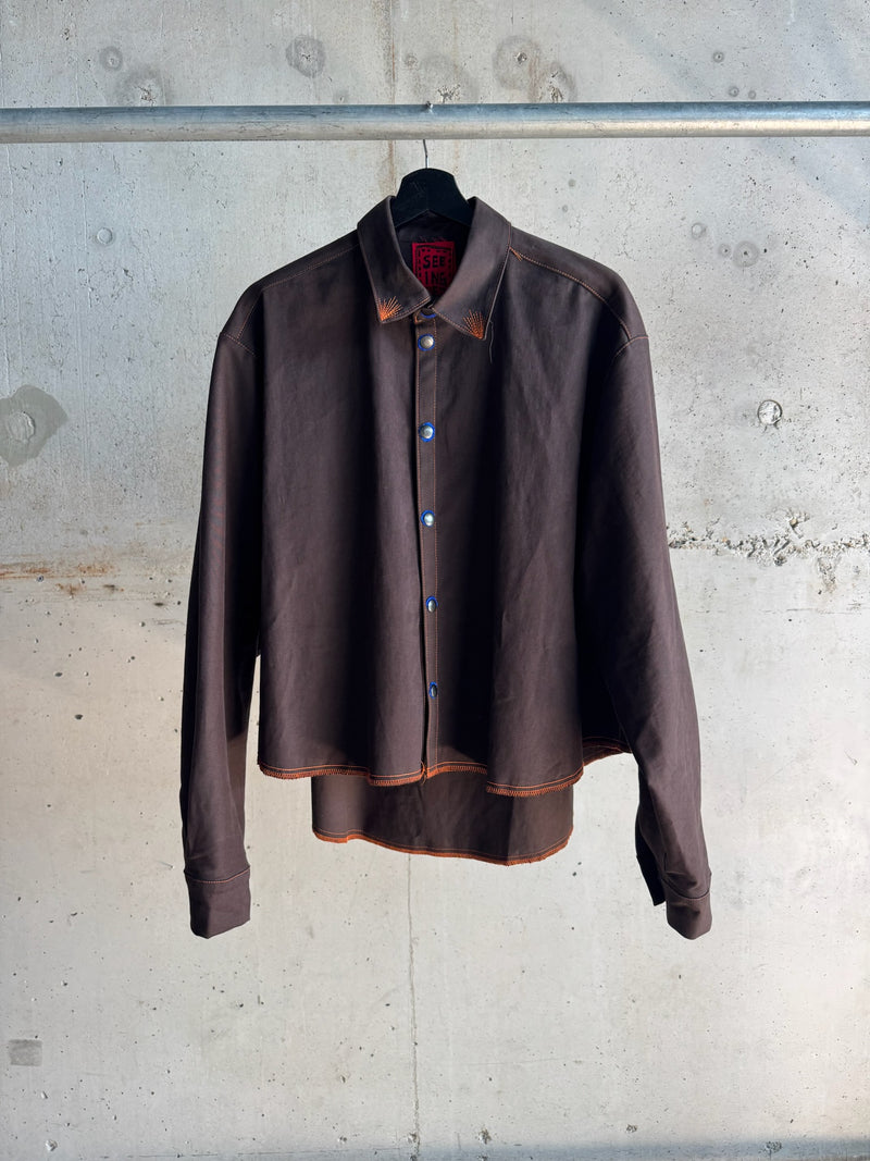 Dark brown long-sleeve button-up shirt with a structured silhouette, featuring contrasting orange topstitching and blue buttons, hanging against a concrete wall background.