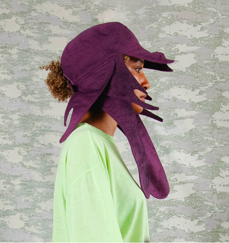 Side profile of model wearing avant-garde deep purple sculptural hat with exaggerated flaps and cutouts, paired with a neon green oversized top – styled against a muted camo background