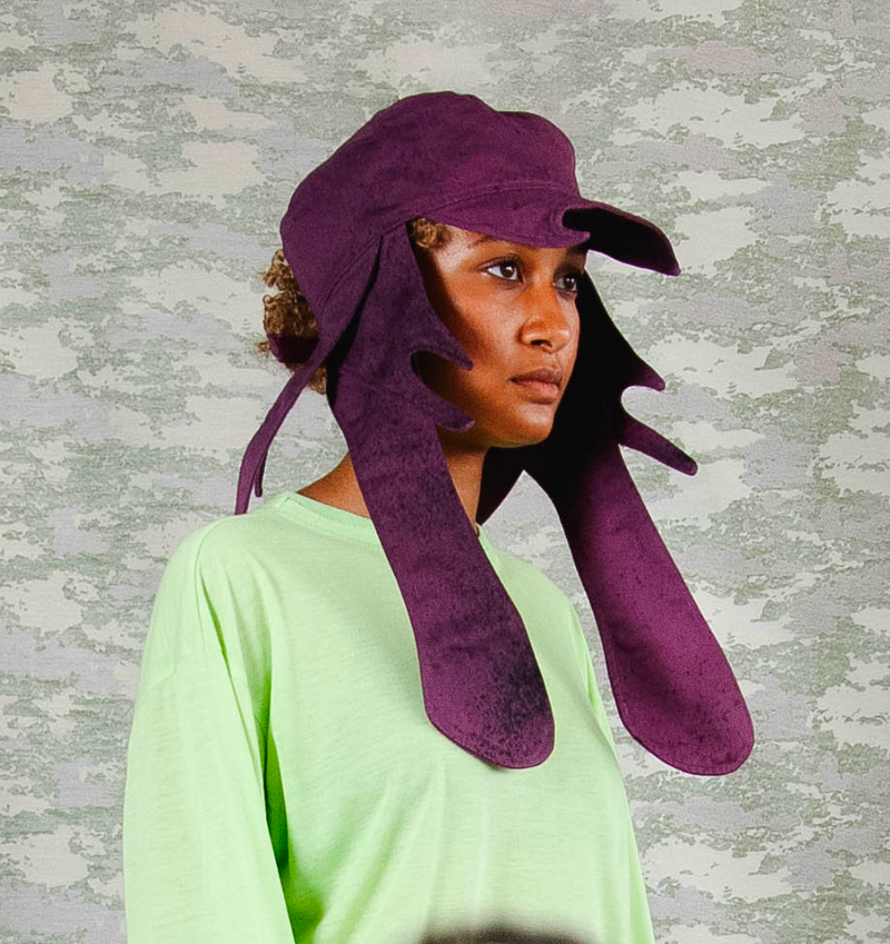 Model wearing sculptural deep purple headpiece with extended abstract flaps, styled with a pastel green oversized top – contemporary avant-garde fashion against a camo-print backgroun